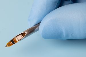 A gloved hand holds a syringe. In the mouth of the syringe sits a small electronic device.