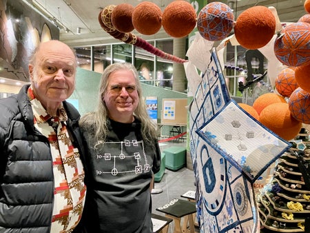 Two men standing next to a large illustrated quilt and a number of large hanging orange basketball-sized spheres.