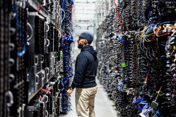 A information halfway worker stands successful a constrictive aisle betwixt gangly server racks packed pinch colorful cables.