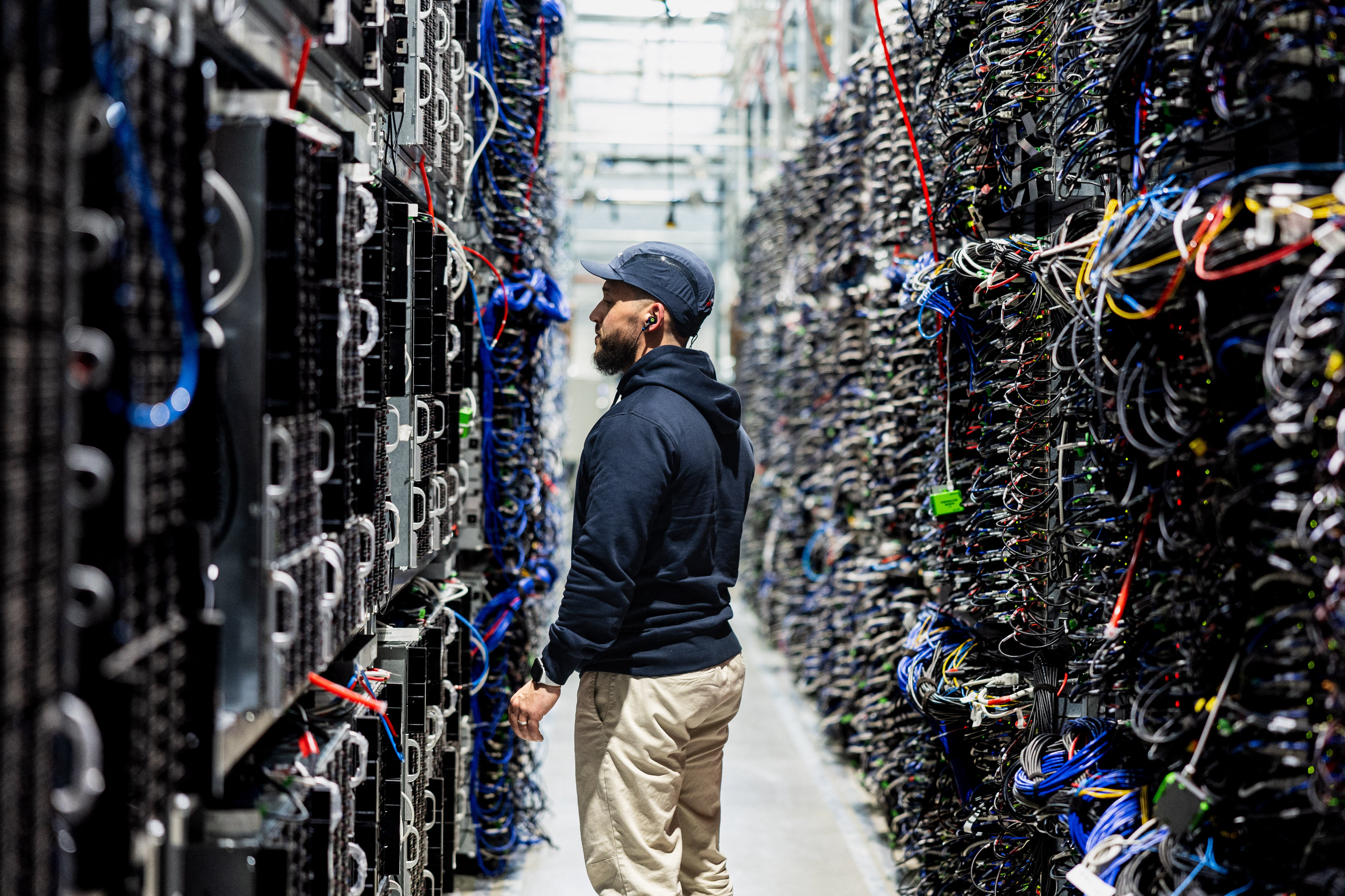 A data center worker stands in a narrow aisle between tall server racks packed with colorful cables.