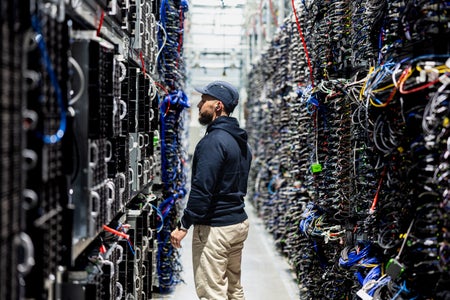 A data center worker stands in a narrow aisle between tall server racks packed with colorful cables.