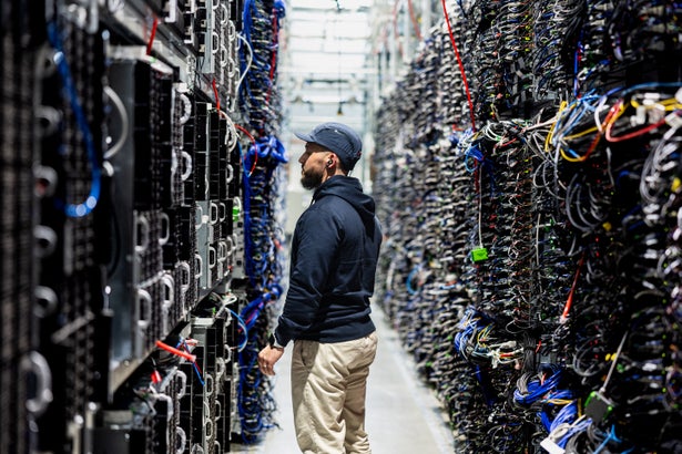 A data center worker stands in a narrow aisle between tall server racks packed with colorful cables.