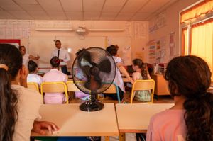 A man in a white dress shirt and tie stands at the front of a classroom full of kids in desks seen from behind. A black fan is on the desk in the foreground.