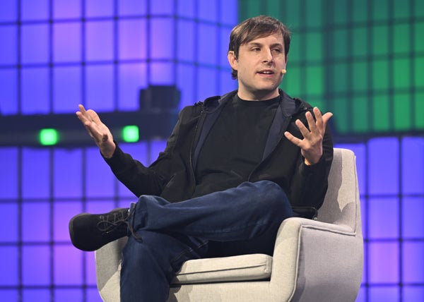 Max Hodak sitting on a stage with his hands out, against a purple and green geometric background.