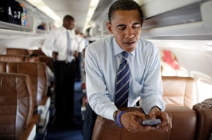 A political figure looks down at a phone while standing in an airplane cabin with aides nearby.