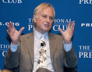 The evolutionary biologist Richard Dawkins speaks while seated on stage in front of a blue background.