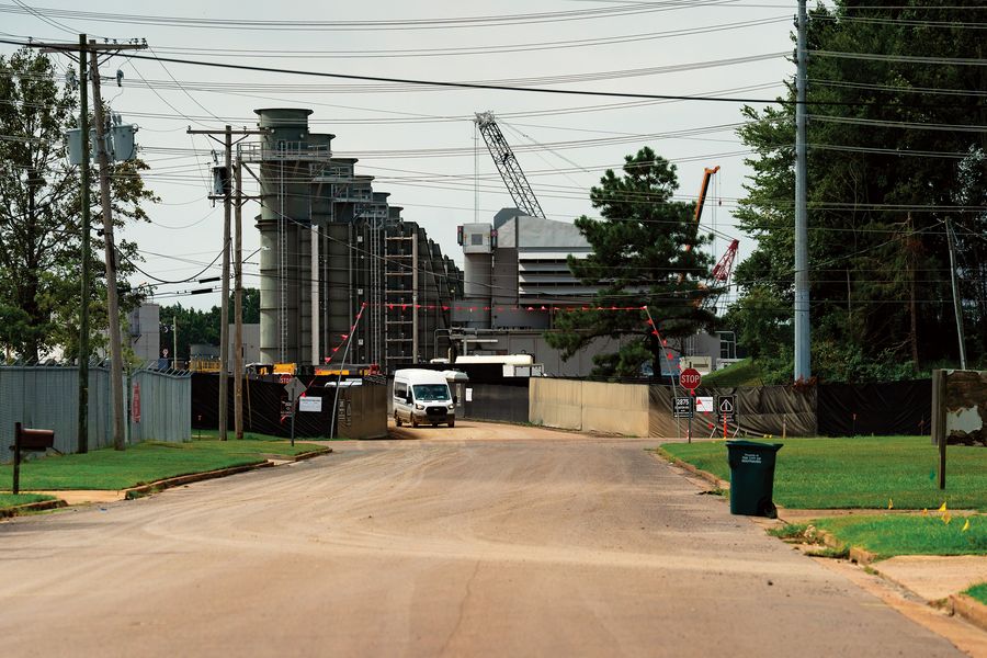 Utility poles, a mailbox and a trash bin line a residential street that ends at tall turbine stacks behind black fencing.