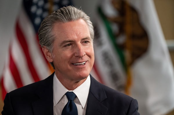 Gavin Newsom, governor of California, in a close-up, with an American flag and a flag of California in the background.
