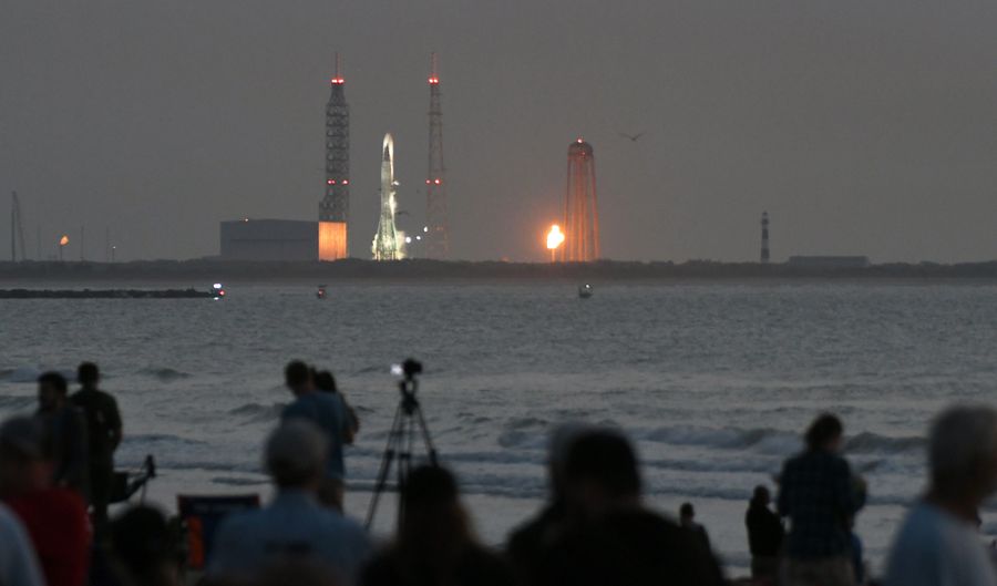 Shadowy spectators in the foreground watch a large rocket on a launch pad in the distance under a dark gray sky.