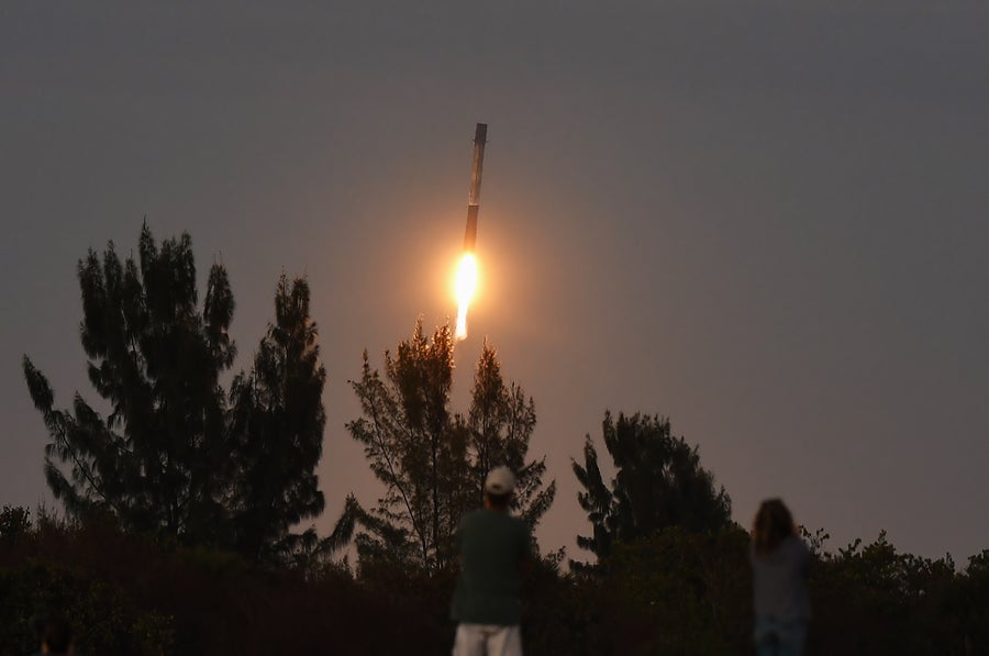A rocket booster descends vertically with engine flame visible as two people watch from below.