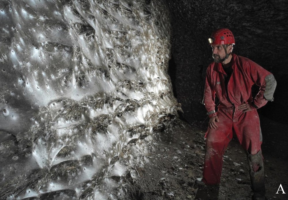 Un espeleólogo con un mono rojo y un casco está a la derecha, su faro ilumina la telaraña gigante en la cueva frente a él.