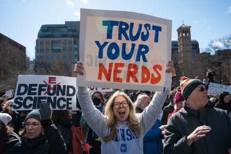 A protest scene focused on a woman holding a sign reading "trust your nerds" in block letters colored blue and red.