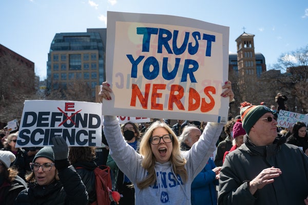 A protest scene focused on a woman holding a sign reading "trust your nerds" in block letters colored blue and red.