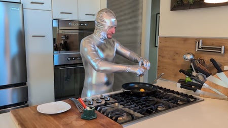 A man wearing a shiny silver full-body suit cracks an egg into a frying pan on a gas stove in a modern kitchen.