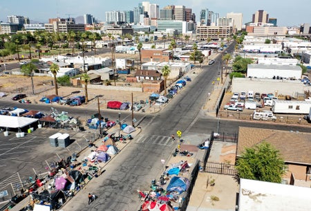 An aerial view shows people walking past a homeless encampment in the afternoon heat with the Phoenix, Arizona skyline in the background