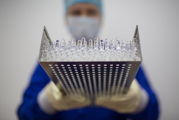 A worker wearing personal protective equipment (PPE) holds a tray containing unlabeled vaccine vials.