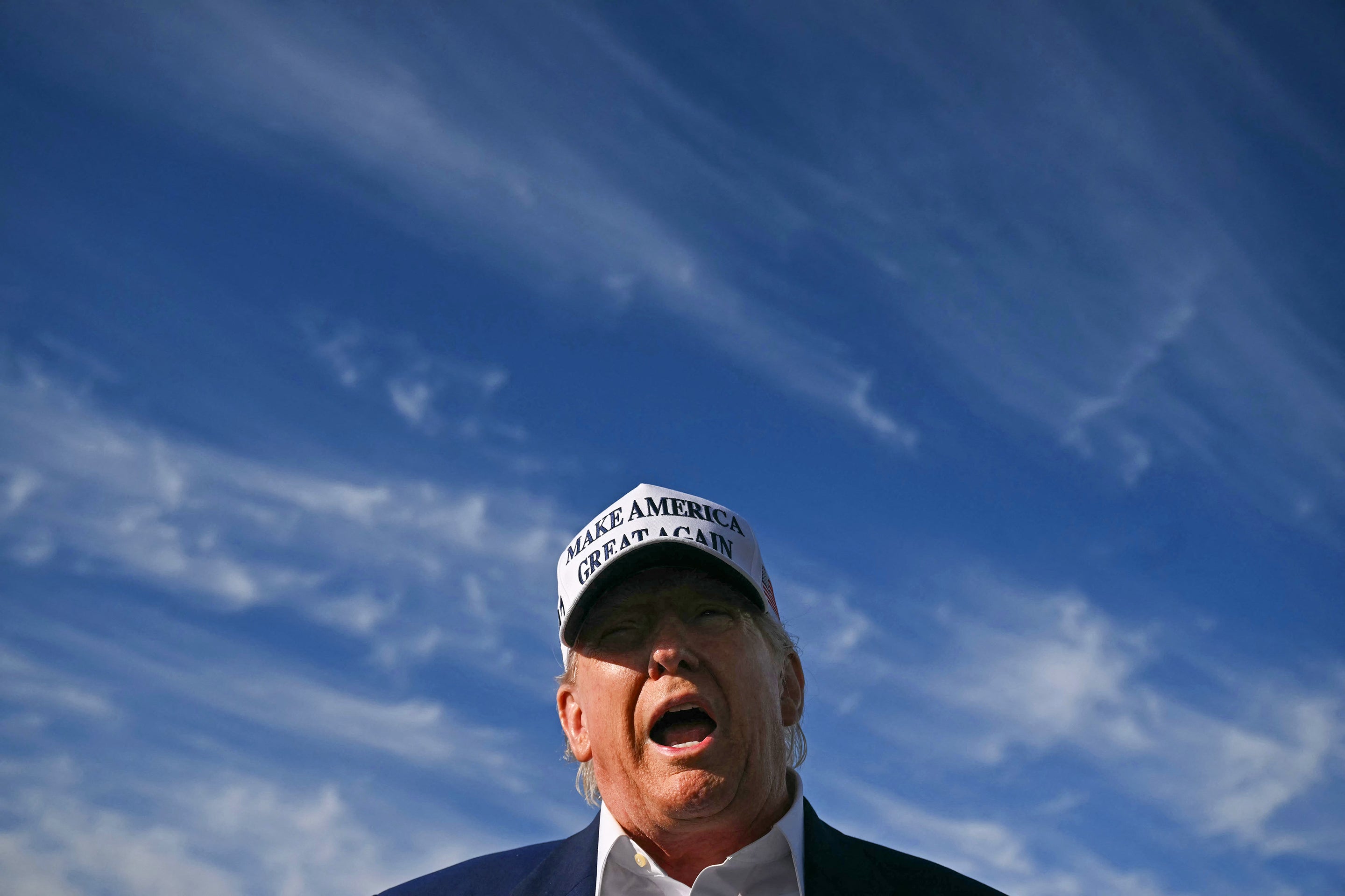 US President Donald Trump speaking to journalists before boarding Air Force One, shot from the shoulders up with blue sky and clouds in the background
