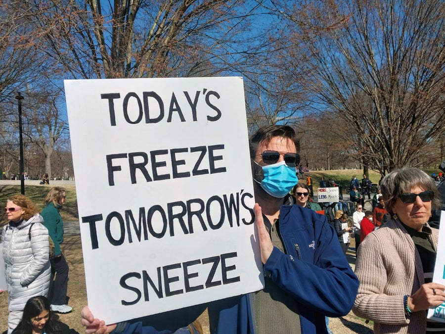 Protester wearing blue face mask holds sign that reads "TODAY'S FREEZE, TOMORROW'S SNEEZE"