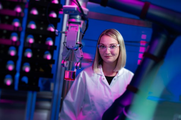 A woman with short blonde hair in a white lab coat and lab glasses in a purple-lit room