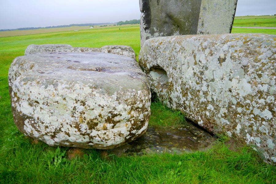 Close-up photograph at Stonehenge showing the Altar Stone partially buried and peeking out from the grass beneath two toppled sarsen stones