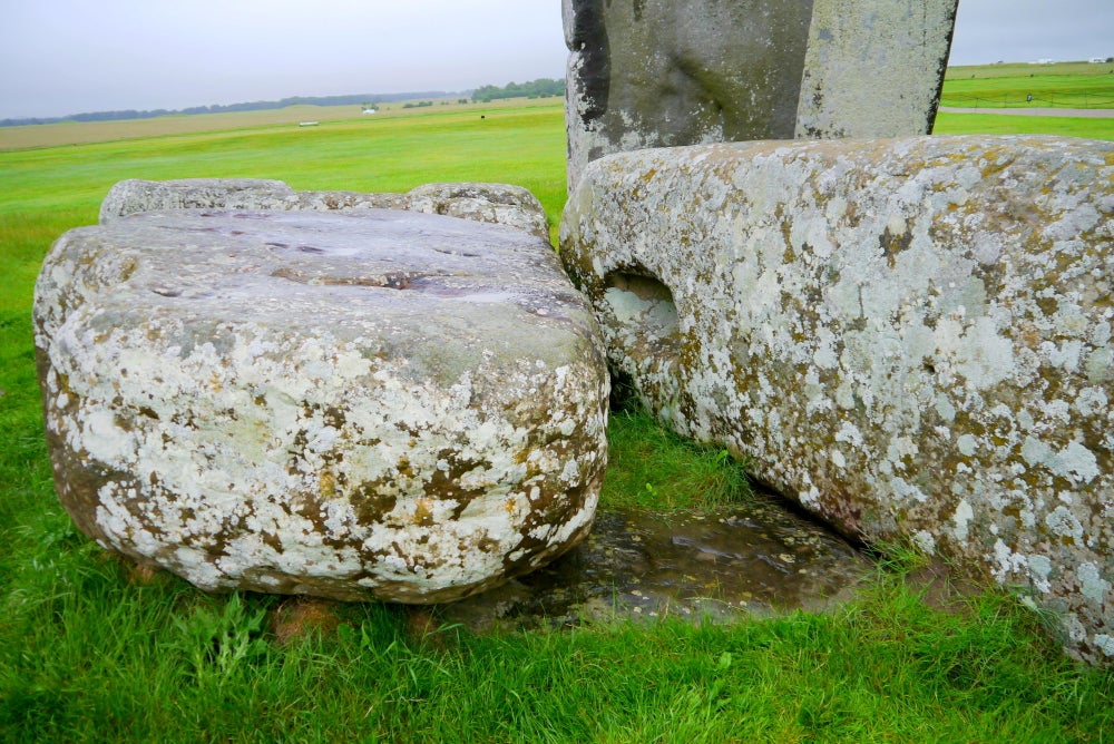 Fotografía de cerca en Stonehenge que muestra la piedra del altar parcialmente enterrada y asomándose entre la hierba debajo de dos piedras Sarsen derribadas