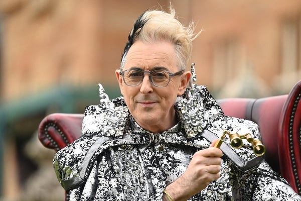 Alan Cumming in a shimmery silver suit, holding a pair of binoculars