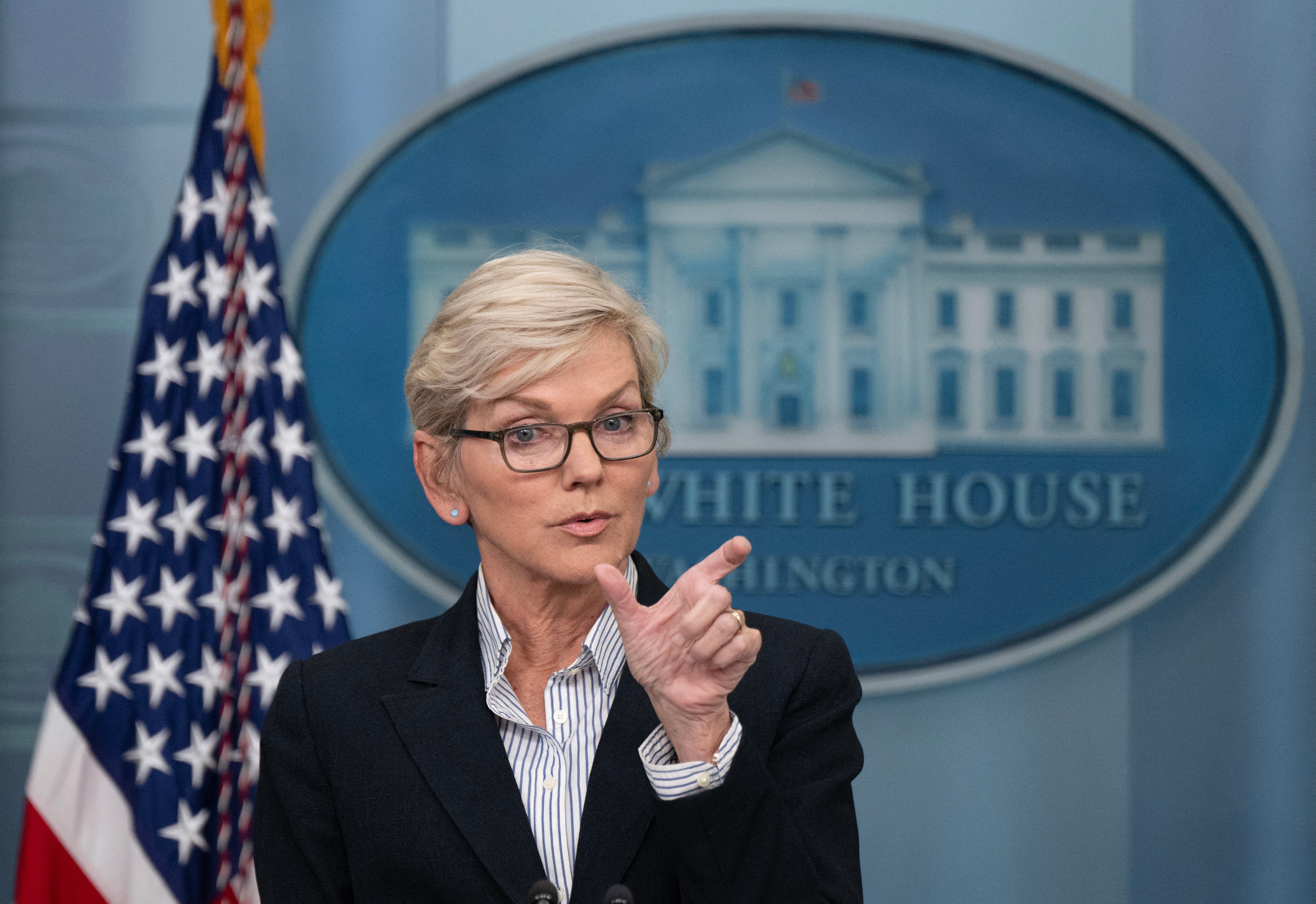 Energy Secretary Jennifer Granholm speaking in front of White House signage and Amercan Flag sizing with fingers.
