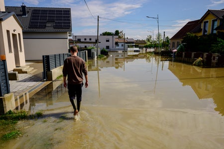 Man in knee high rain boots walks down a flooded residential street