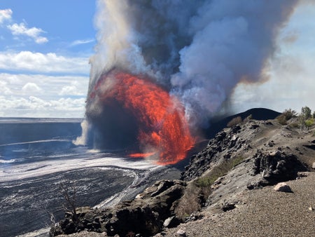 Orange lava and gray rocks and ash spew from the brown and gray ground against a blue sky with white clouds