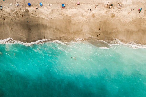Direct aerial overhead view of a vibrant teal ocean, sand beach and colorful beach umbrellas.