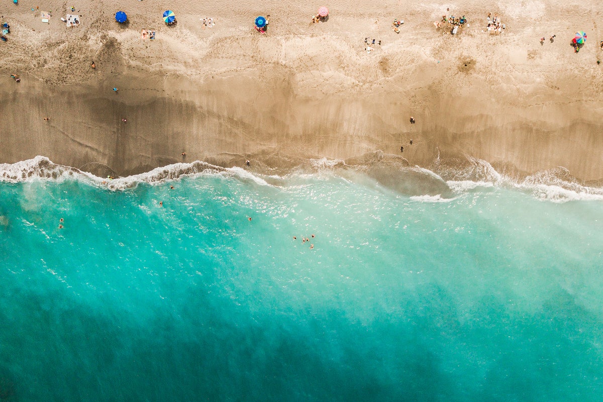 Direct aerial overhead view of a vibrant teal ocean, sand beach and colorful beach umbrellas.