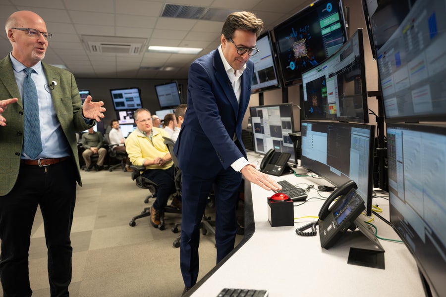 A man in a suit presses a large red button while several onlookers applaud, in a control room festooned with monitors and computers.