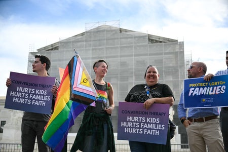 Protestors holding signs and a Pride flag standing in front of the columned Supreme Court building covered by scaffolding.