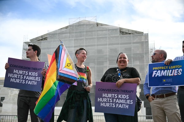 Protestors holding signs and a Pride flag standing in front of the columned Supreme Court building covered by scaffolding.
