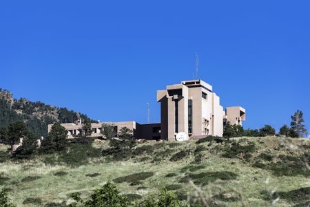 A blocky beige building atop a green hill with a blue sky in the background