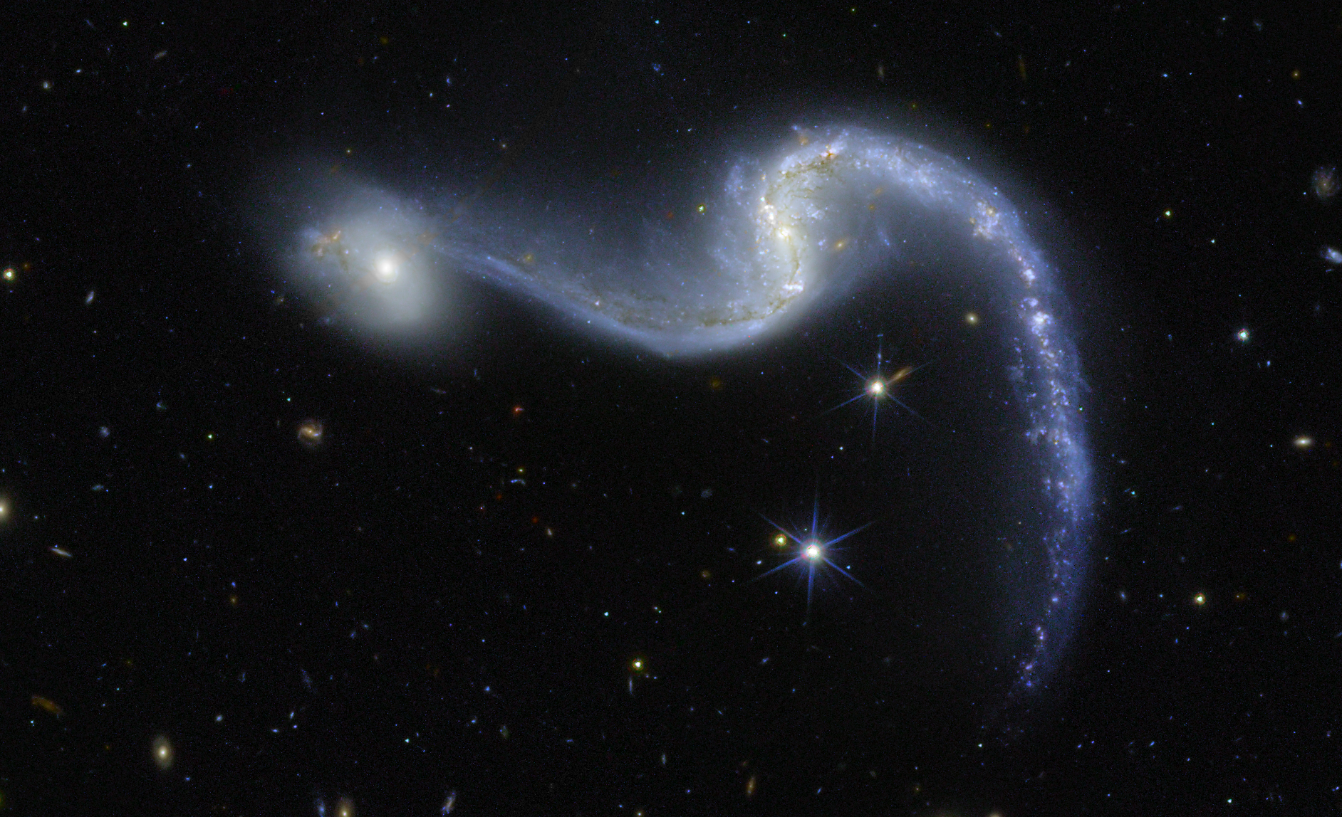 An image of space made by the Euclid telescope shows a bright barred spiral galaxy with two sweeping arms glowing in shades of blue and white against a deep black background dotted with stars. Its core is luminous. Its spiral arms curve gracefully outward to the left and down to the right, resembling a cosmic garland draped across the scene. A smaller, faint and round galaxy appears to the left of the barred spiral galaxy.