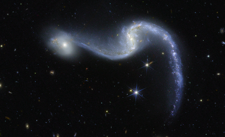 An image of space made by the Euclid telescope shows a bright barred spiral galaxy with two sweeping arms glowing in shades of blue and white against a deep black background dotted with stars. Its core is luminous. Its spiral arms curve gracefully outward to the left and down to the right, resembling a cosmic garland draped across the scene. A smaller, faint and round galaxy appears to the left of the barred spiral galaxy.