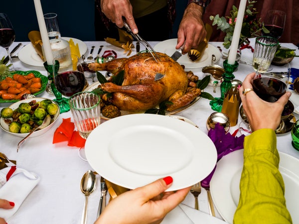 over the shoulder view of a woman holding an empty plate out to a man carving and serving a holiday turkey at dinner table