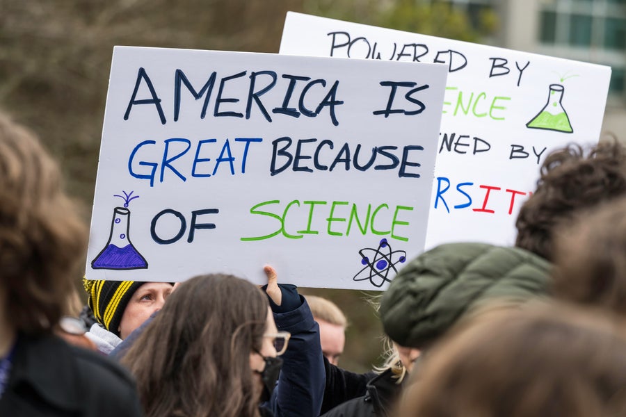 Participants attending the Hands Off Our Healthcare, Research and Jobs rally at University of Washington Portage Bay Campus in Seattle hold signs. In the foreground a sign reads, "America is great because of science."