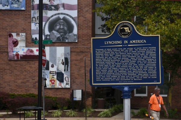 A historic marker detailing "Lynching in America" is seen on a street in Annapolis, Maralynd with street art installed on a building's wall in the background as an unidentified man walks by on the sidewalk