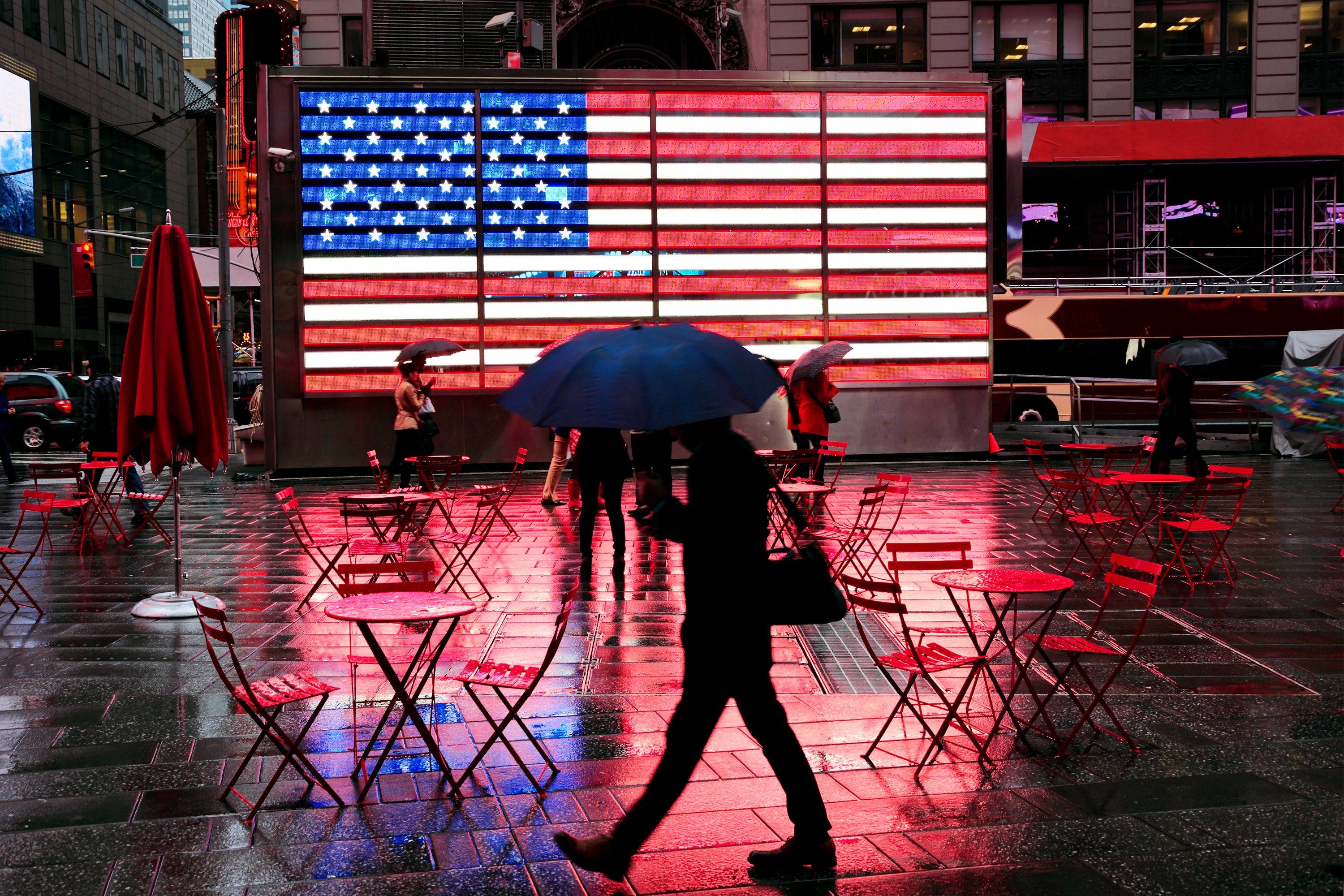 Pedestrian carrying umbrella and shoulder bag silhouette against the famous illuminated American Flag in very wet, dark Times Square