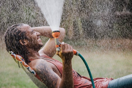 A man sitting outside, spraying a hose up, with water falling on him.