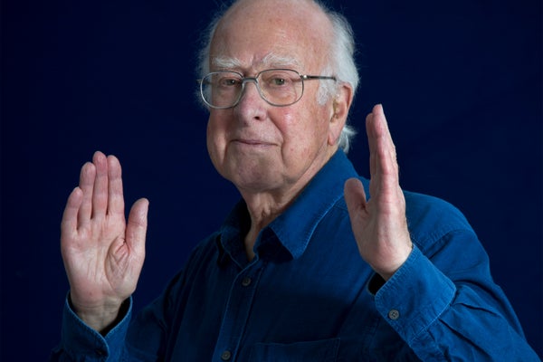 Portrait of Peter Higgs in blue shirt and glasses both hands and apart.