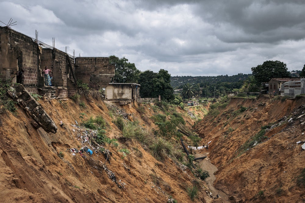 Una vista por un barranco urbano en Kinshasa, DRC. A la izquierda en el borde superior del barranco, una mujer y un niño se paran en parte de una casa que se derrumbó en el barranco
