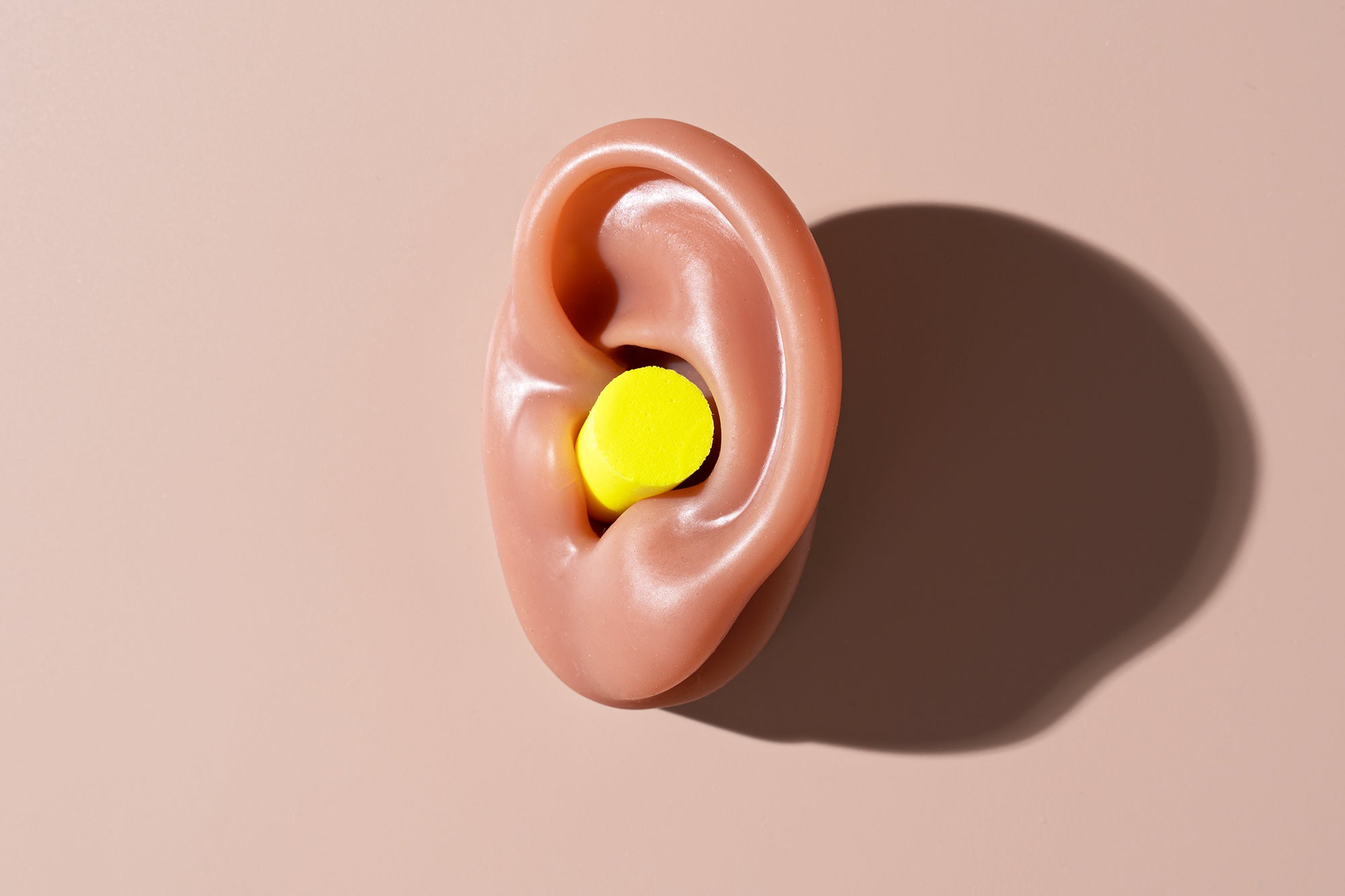 Yellow colored, traditional sound insulation earplug inserted in a 3-D model of a human ear on a brown surface with high-key lighting and a sharp, dark shadow