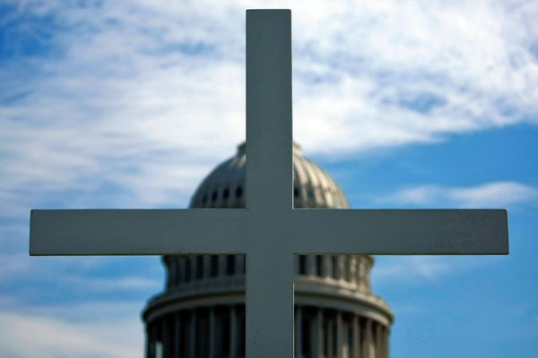 Close up photograph of a 16-foot cross with the dome of the U.S. Capitol Building in Washington D.C. visible, out of focus, behind