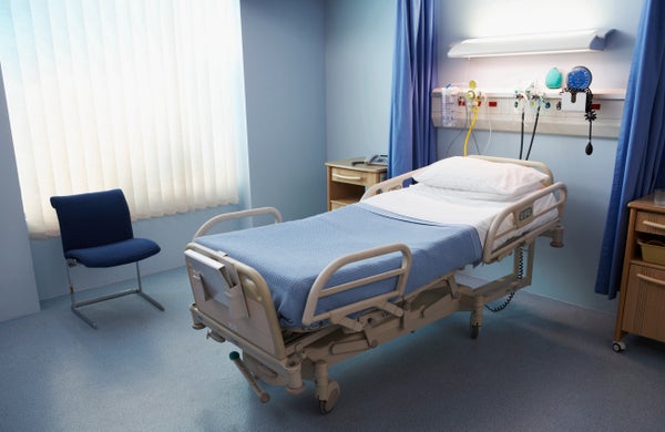 Empty Hospital Bed in a Ward with blue curtains and covers and chair.