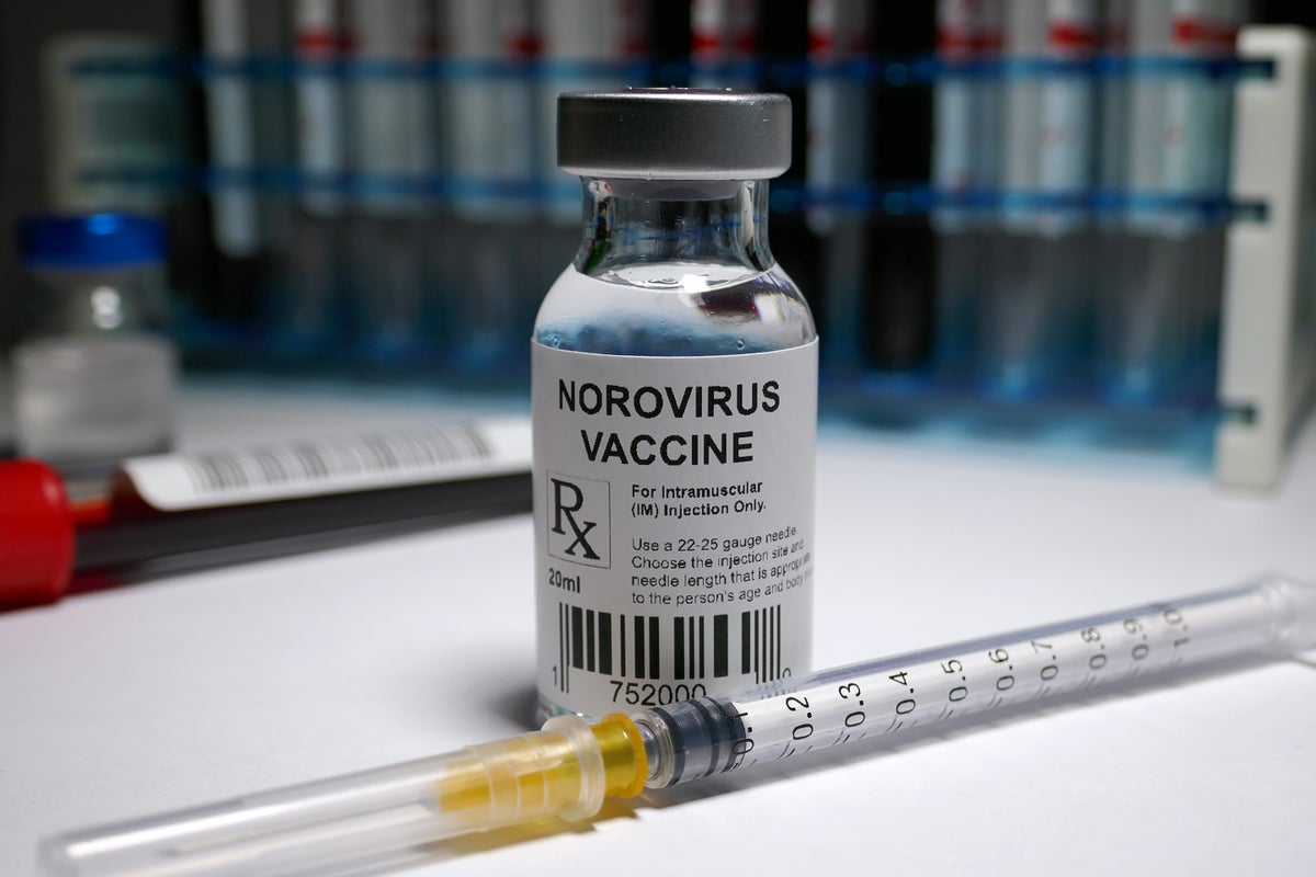 Photo illustration of a norovirus vaccine in a clear glass vial standing on a white surface with an injection needle laying in front of the vial