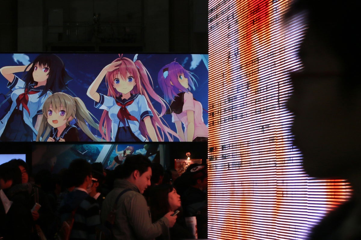 People visit the venue of AnimeJapan with female anime characters on a screen and a male silhouette of a spectator on the right.