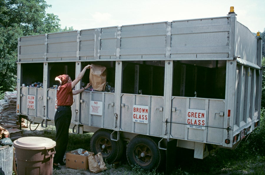 A woman wearing a red bandana in front of a grey recycling container sorting materials.